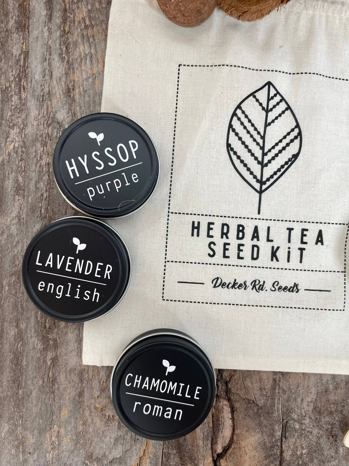 Three black lids labeled 'Hyssop', 'Lavender', and 'Chamomile' on a wooden surface with a 'Herbal Tea Seed Kit' bag.