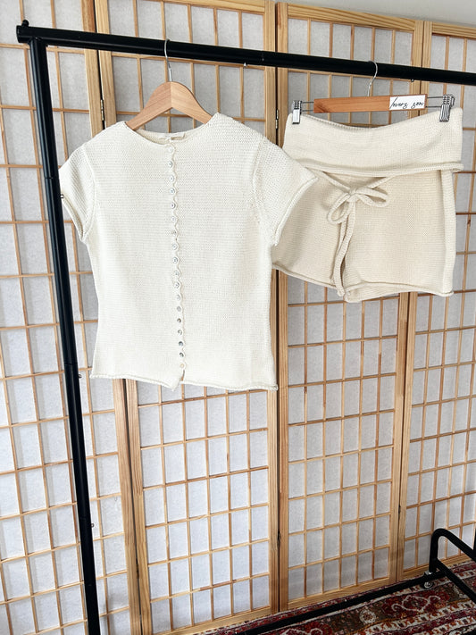 An off-white short-sleeve textured knit top with a button-down front and matching off-white shorts with a tie waist, displayed on wooden hangers on a clothing rack in front of a Japanese-style folding screen.