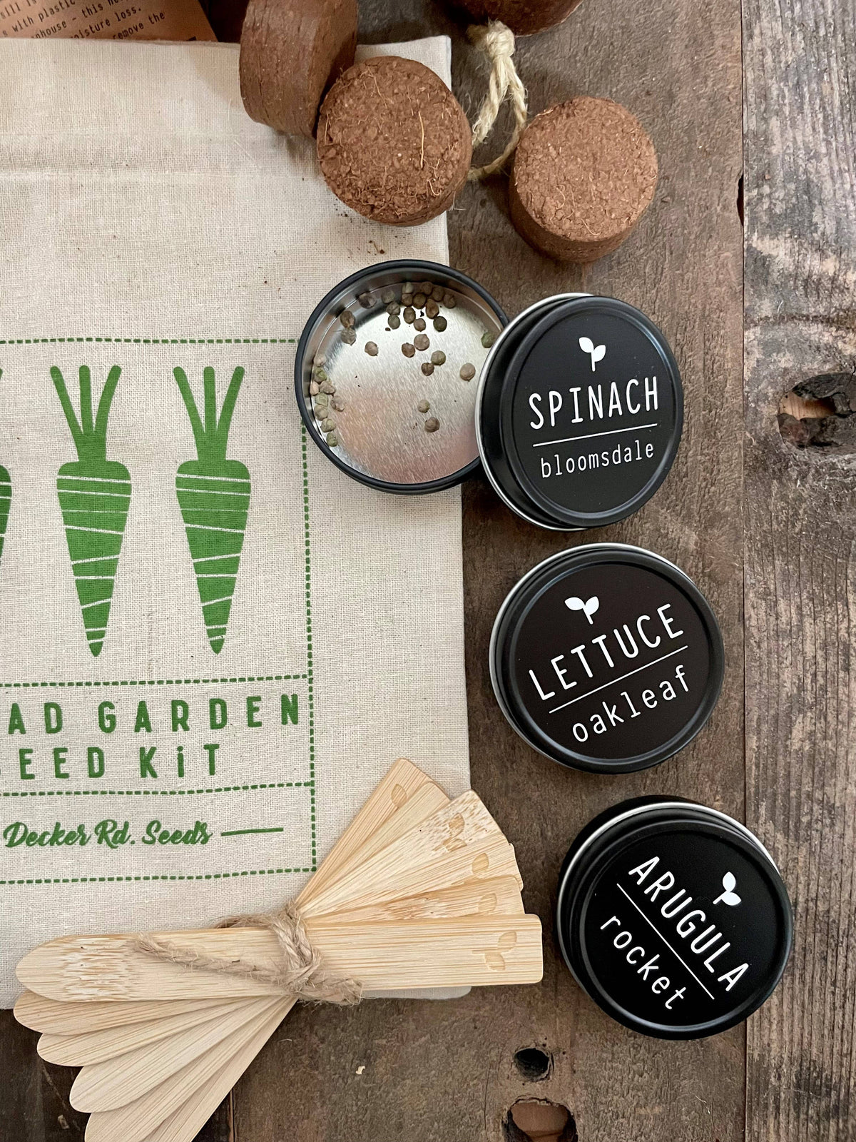 Seedling pots labeled 'Spinach', 'Lettuce', and 'Arugula' next to a seed packet on a wooden surface.