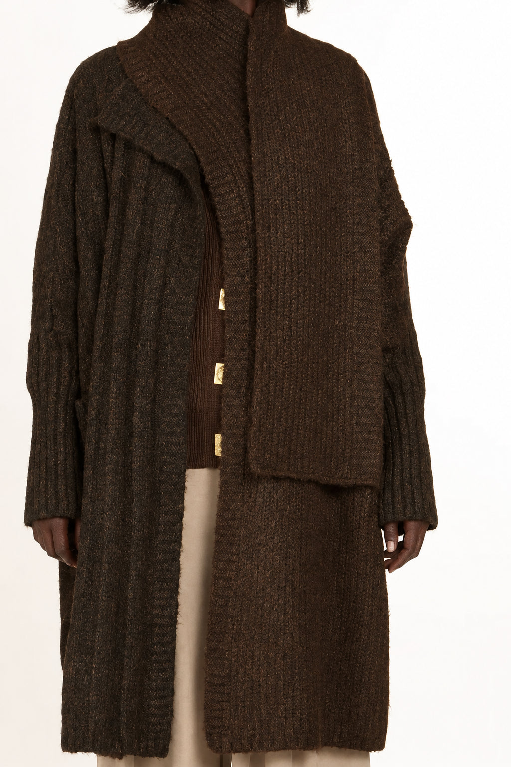 A woman stands against a plain light background wearing a dark brown textured knit cardigan with a high collar and gold-tone buttons, layered over a brown top and beige wide-leg pants.