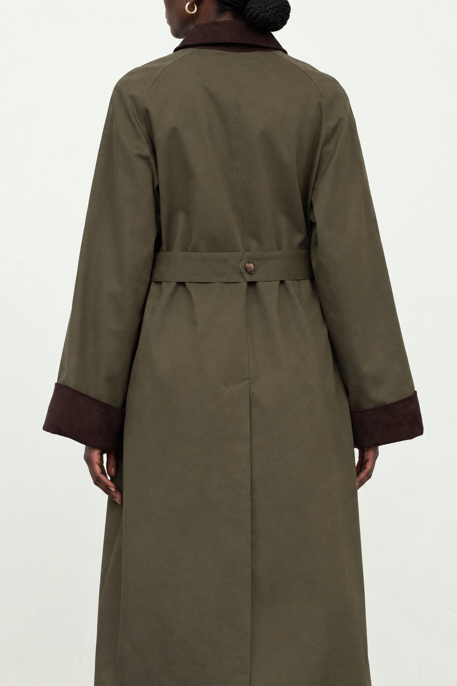 A person shown from the back wearing a long olive green trench coat with dark brown trim at the collar and cuffs, photographed against a plain light background.