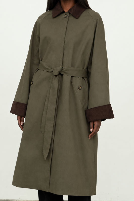 An olive green trench coat with dark brown contrast collar, cuffs, and belt, shown on a model against a plain white background.