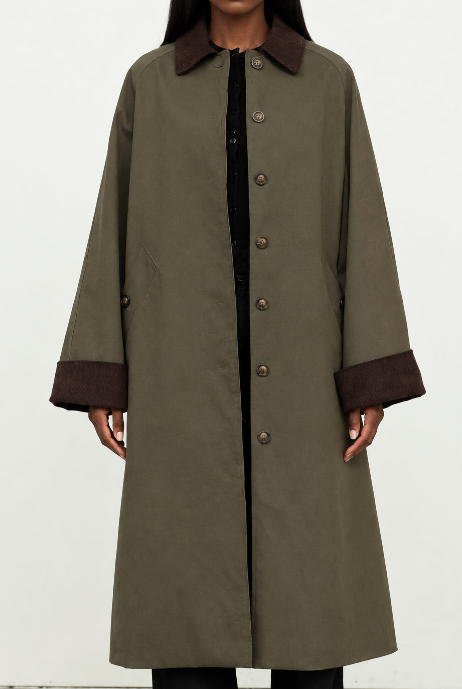 A long olive green coat with dark brown collar, cuffs, and button details, worn by a model over a black top against a plain off-white background.