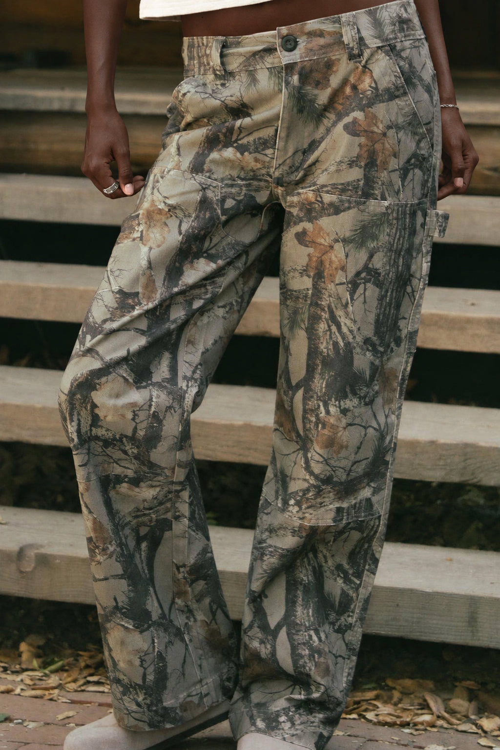 A woman stands on wooden steps in front of a log cabin wearing a white cropped graphic T-shirt and camouflage wide-leg pants.