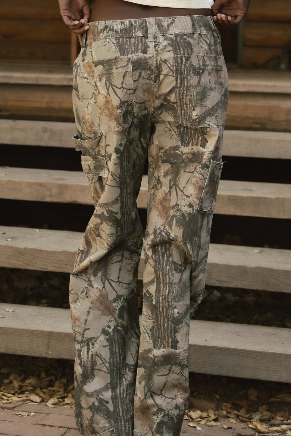 A woman stands with her back to the camera wearing camouflage cargo pants and a white cropped t-shirt, posing in front of a rustic wooden cabin with steps.