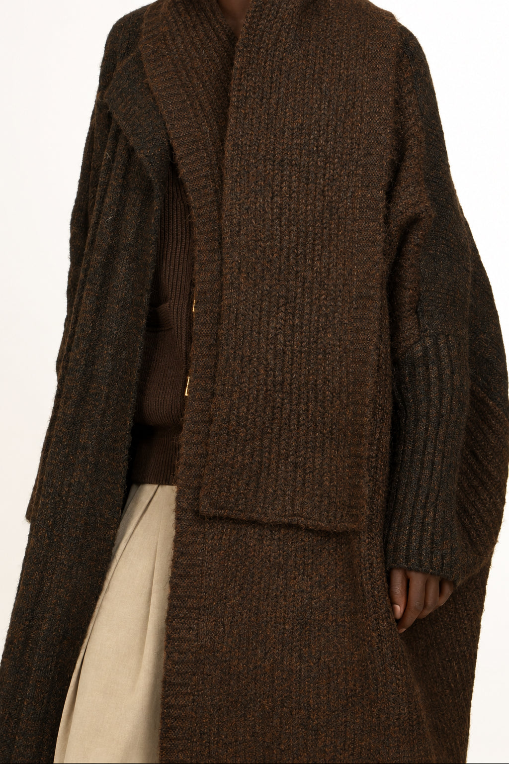 A woman wearing a dark brown chunky knit wrap coat over a brown top and beige skirt, photographed against a plain white studio background.