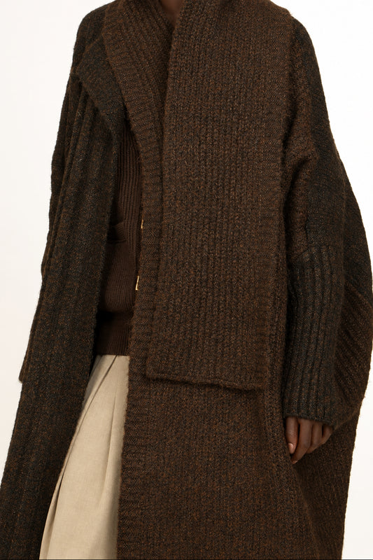 A woman wearing a dark brown chunky knit wrap coat over a brown top and beige skirt, photographed against a plain white studio background.