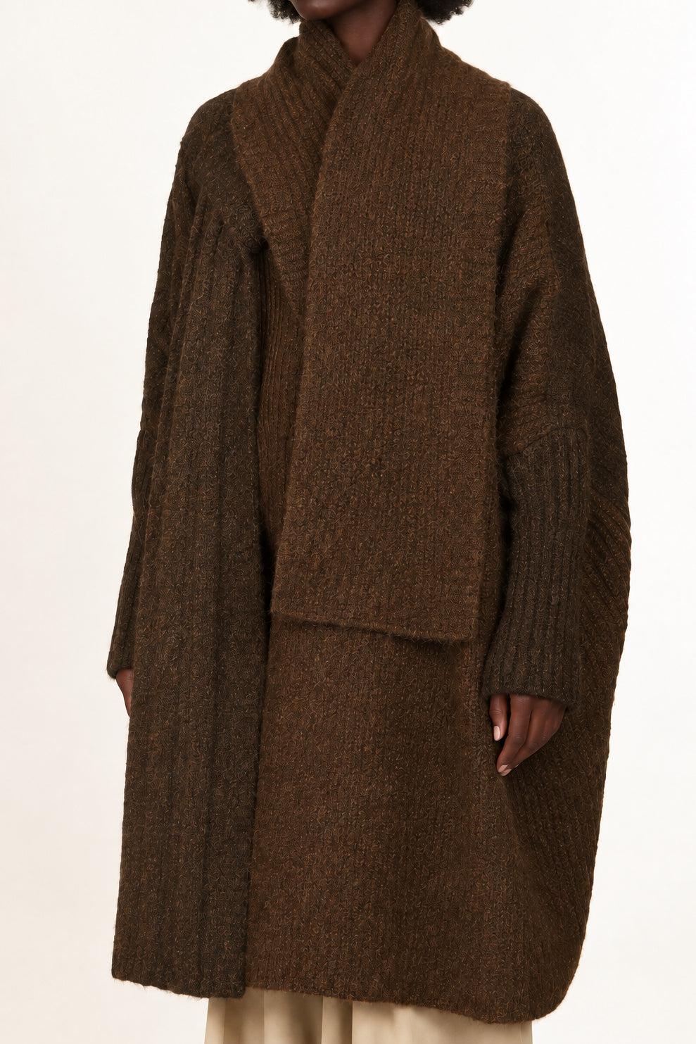 A model wearing a dark brown textured knit wrap coat over a matching brown top and beige wide-leg trousers, photographed against a plain white studio background.
